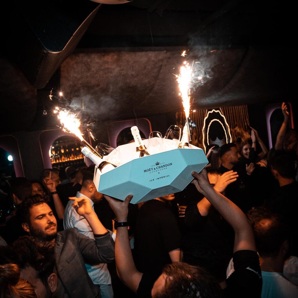 Eine Gruppe von Clubgästen feiert ausgelassen, während eine Person ein Tablett mit Moët & Chandon Champagnerflaschen und brennenden Wunderkerzen hochhält. Die lebhafte Atmosphäre im Club wird durch die jubelnden Gäste und die funkelnden Lichter verstärkt.